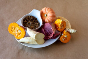 Prato com ingredientes naturais, como abóbora, carne vermelha e mandioca, rodeando uma vasilha de ração.